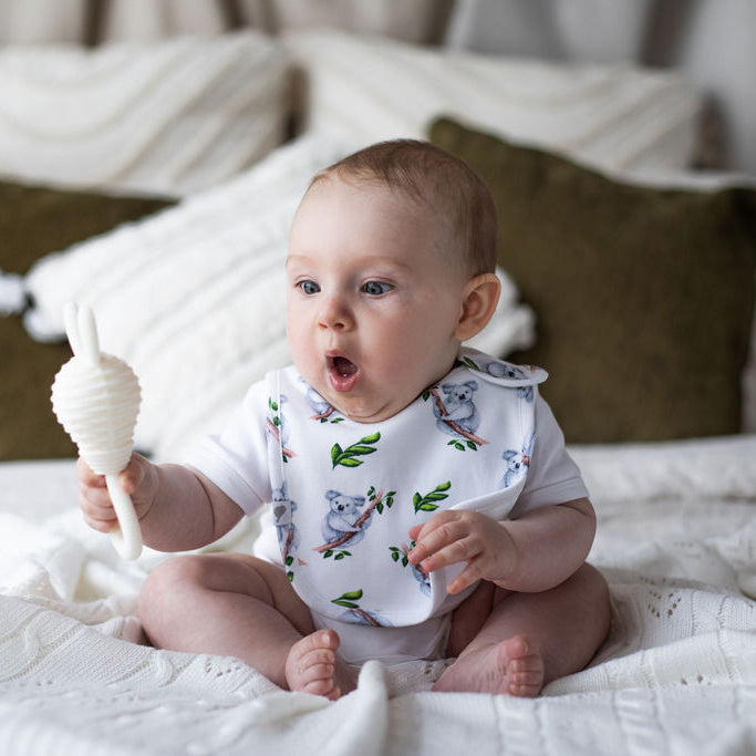 Baby sitting on a bed holding a rattle, wearing a bib with animal prints.