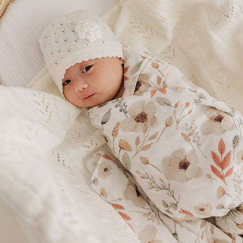 Baby swaddled in a Boho muslin with a cream flower knit hat, lying on a cream fern blanket.