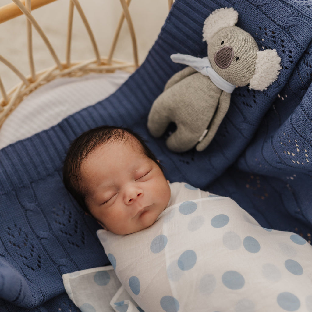 Newborn baby swaddled in a blue spot muslin with a soft knitted koa koala rattle on a denim fern blanket.