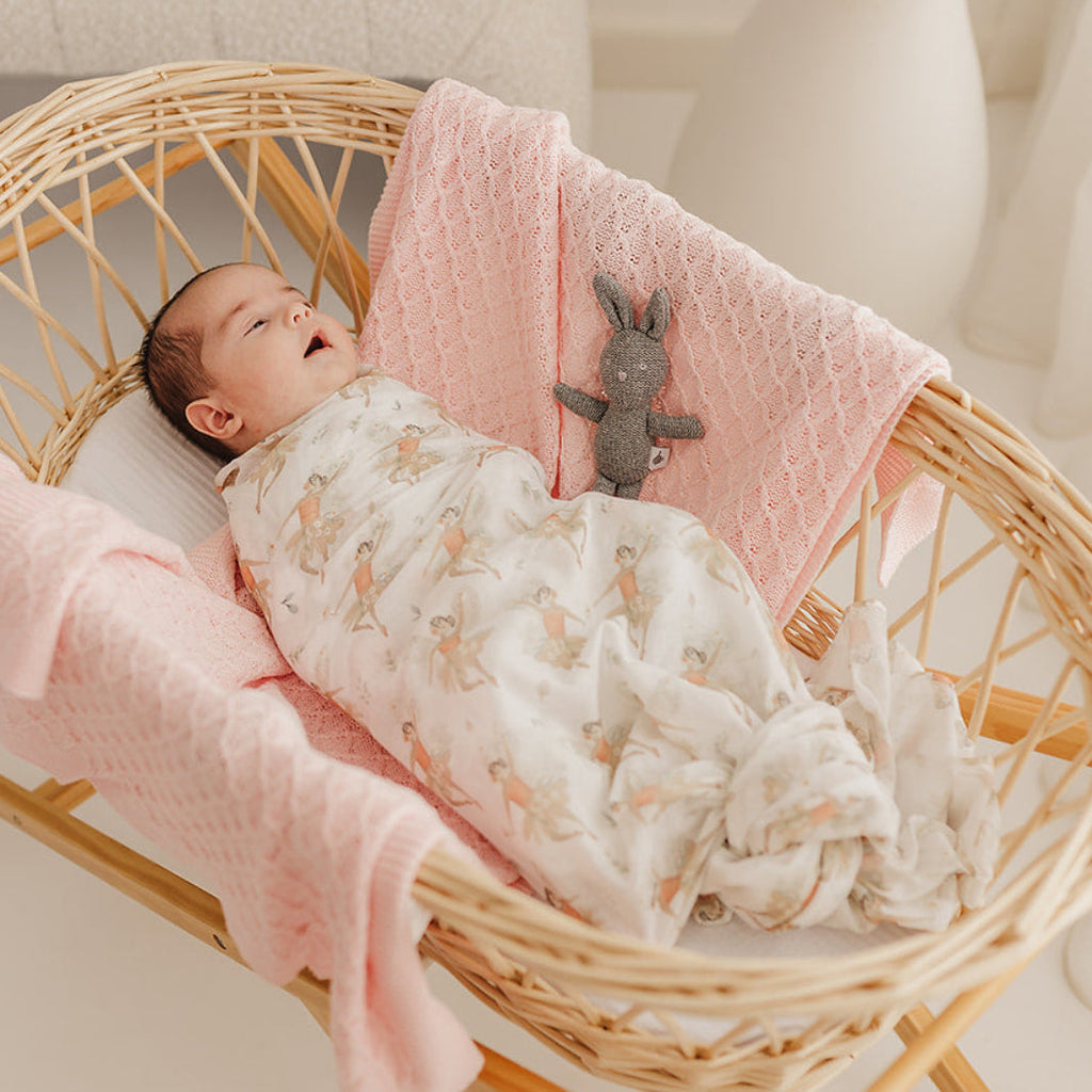 Newborn baby swaddled in a fairy muslin lying in a moses basket with pink lace blanket and grey knitted bunny rattle, surrounded by dried plants.
