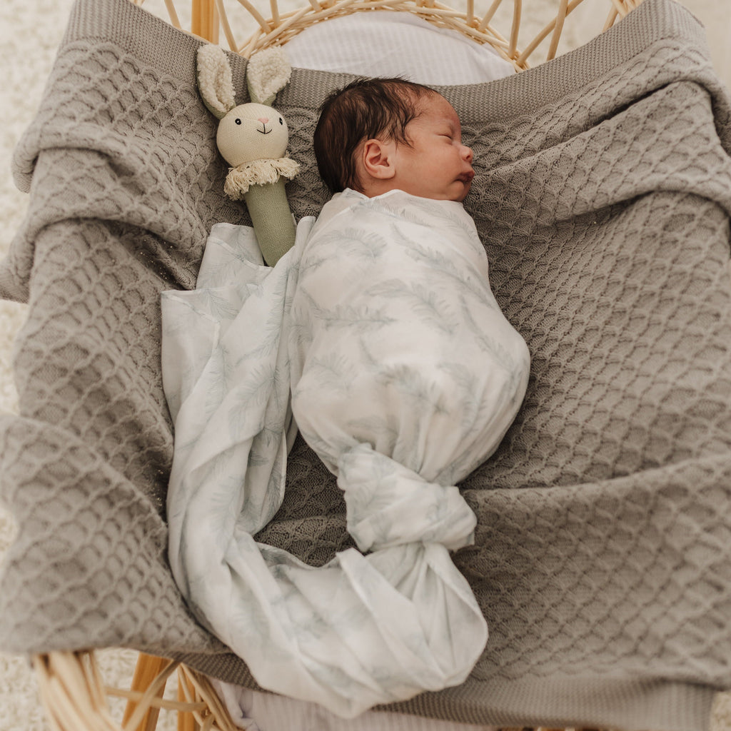 Newborn baby wrapped in feather muslin lying on a grey lace blanket with a sage bunny rattle in a wicker basket.
